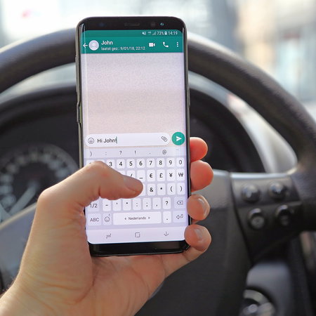 Driver holding a smartphone with WhatsApp open behind the wheel of a car while stopped in traffic.