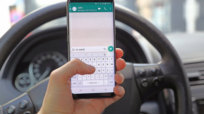 Driver holding a smartphone with WhatsApp open behind the wheel of a car while stopped in traffic.