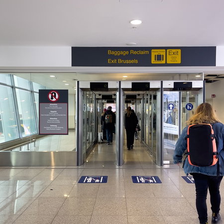 Travellers walk through an arrivals corridor at Brussels Airport beneath exit and baggage reclaim signs.
