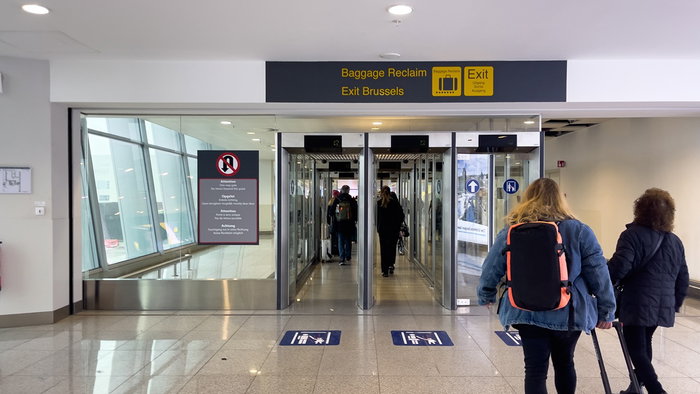 Travellers walk through an arrivals corridor at Brussels Airport beneath exit and baggage reclaim signs.