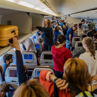 Busy interior of a plane cabin while person retrieves cabin bag from overhead bin.