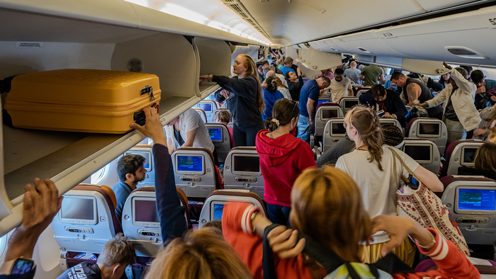 Busy interior of a plane cabin while person retrieves cabin bag from overhead bin.