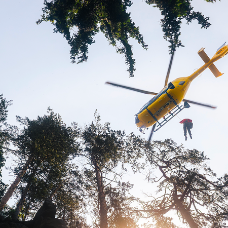 Mountain rescue helicopters lowers man between trees.