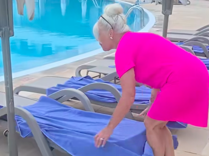 Woman in bright pink dress removes towels from sunbeds by a pool