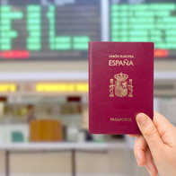 Hand holding a Spanish passport in front of an airport departures board.