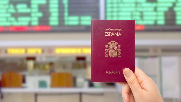 Hand holding a Spanish passport in front of an airport departures board.