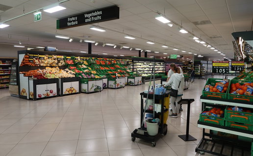 Inside a Spanish supermarket fruit and vegetable section, with fresh produce displays and bilingual signage above the aisles.