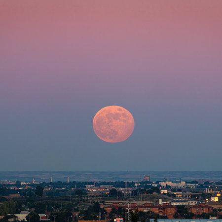 The Strawberry Moon rises above the horizon at dusk, casting a warm orange glow over a city skyline.