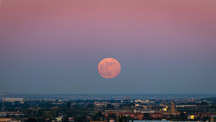 The Strawberry Moon rises above the horizon at dusk, casting a warm orange glow over a city skyline.