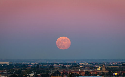 The Strawberry Moon rises above the horizon at dusk, casting a warm orange glow over a city skyline.