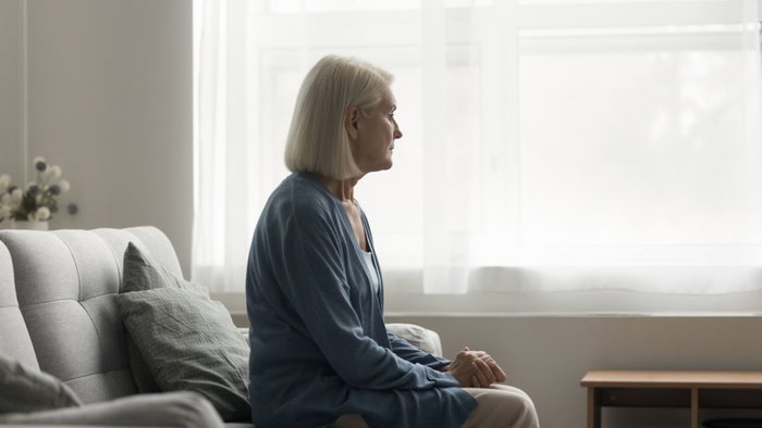 Side profile of a pensive elderly woman with grey hair sitting alone on a couch and looking out a bright window, representing memory loss or dementia.