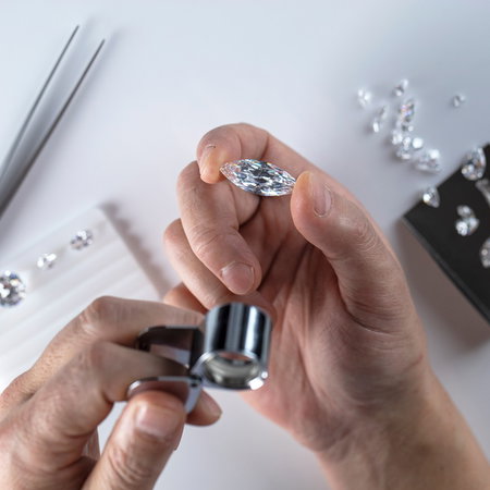 A gem expert examines a loose diamond with a loupe beside other stones on a white table.