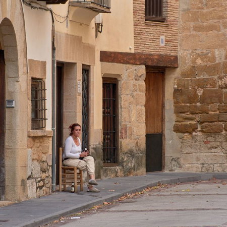A woman sitting on a wooden chair outside a traditional stone house on a quiet street in a rural Spanish village.