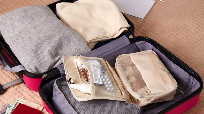 An open suitcase packed with clothes, organiser cubes, and a clear travel first aid pouch filled with blister packs of pills, capsules, and a spray bottle.