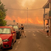 Cars parked on a sidewalk and onlookers watch flames approach a village