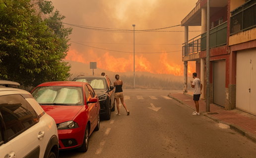 Cars parked on a sidewalk and onlookers watch flames approach a village