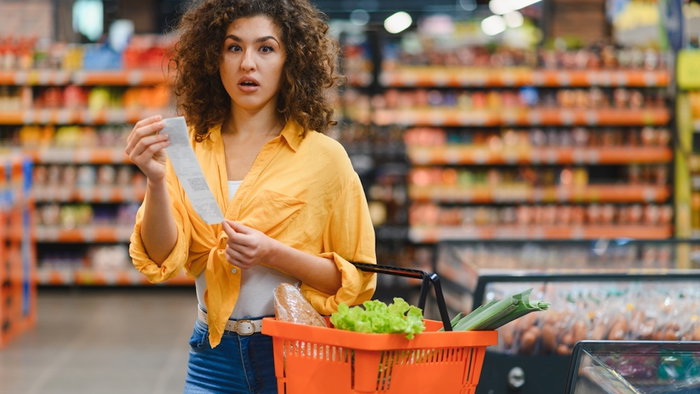 Woman holding a long receipt with a surprised expression in a grocery supermarket aisle, reflecting high food prices