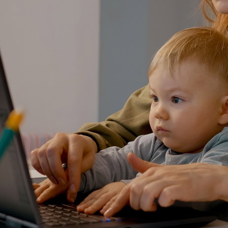 A mother works on a laptop at home while holding a small child on her lap.