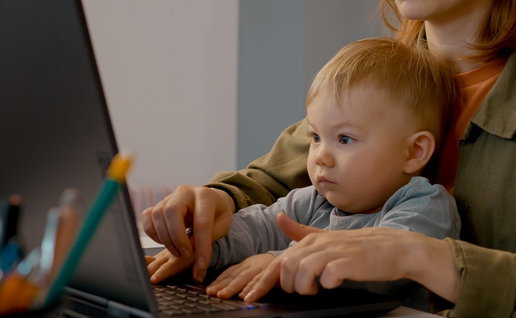 A mother works on a laptop at home while holding a small child on her lap.