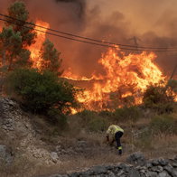 Firefighter in yellow protective gear bent over on a rocky hillside, working near a massive, raging wildfire consuming trees and hills under a thick orange smoke-filled sky