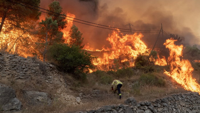 Firefighter in yellow protective gear bent over on a rocky hillside, working near a massive, raging wildfire consuming trees and hills under a thick orange smoke-filled sky