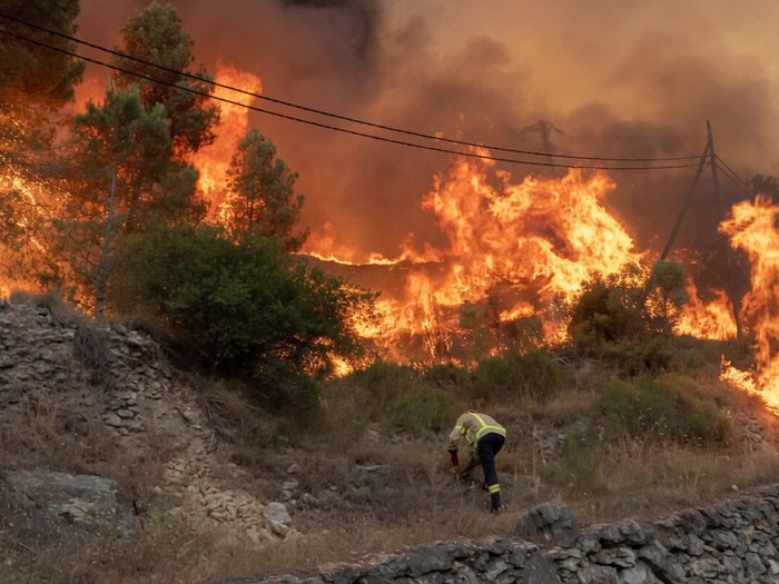 Firefighter in yellow protective gear bent over on a rocky hillside, working near a massive, raging wildfire consuming trees and hills under a thick orange smoke-filled sky