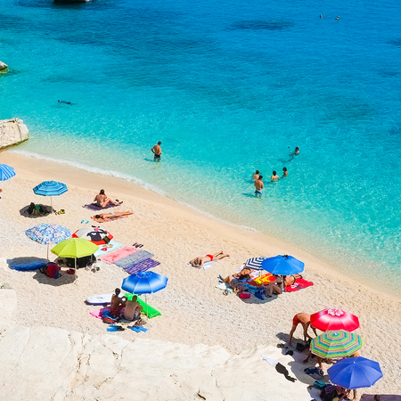 Holidaymakers swimming and relaxing beneath colourful umbrellas at Cala Goloritzé beach in Sardinia, Italy.