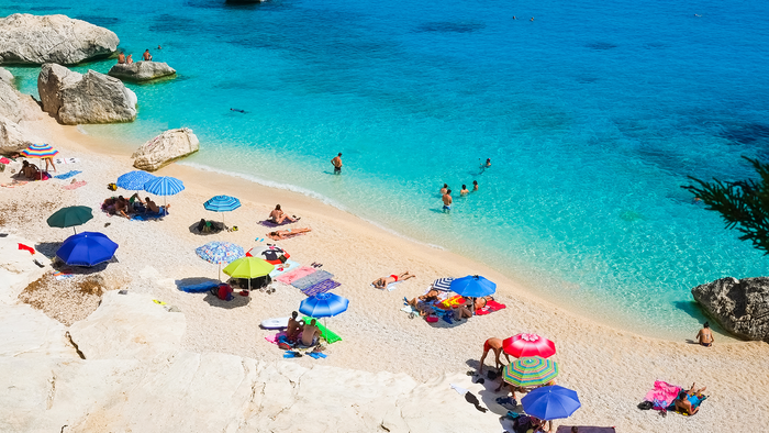 Holidaymakers swimming and relaxing beneath colourful umbrellas at Cala Goloritzé beach in Sardinia, Italy.