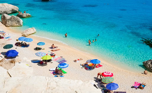 Holidaymakers swimming and relaxing beneath colourful umbrellas at Cala Goloritzé beach in Sardinia, Italy.