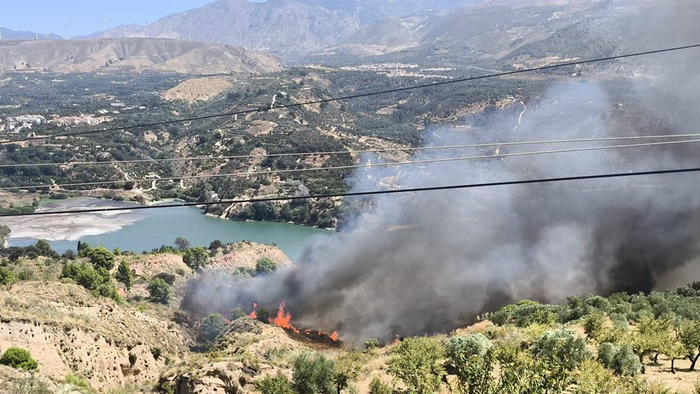 Flames and thick black smoke rising from hillside vegetation beside the Béznar reservoir.