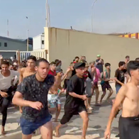 A large group of mostly young men, some shirtless and wearing wet clothing, running along a road beside border facilities in Ceuta.