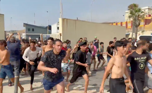 A large group of mostly young men, some shirtless and wearing wet clothing, running along a road beside border facilities in Ceuta.