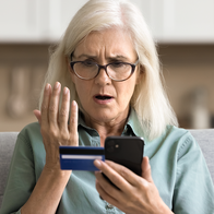 Older woman holding a bank card and looking worried at her phone.