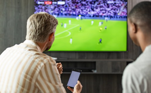 Two friends watching a football game on TV one is holding a smartphone going to place a bet