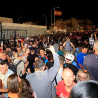 Large crowds pass through the Gibraltar-La Línea border crossing at night, with people filming on phones and a Spanish flag flying above.