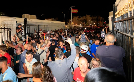 Large crowds pass through the Gibraltar-La Línea border crossing at night, with people filming on phones and a Spanish flag flying above.