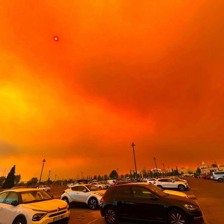 An Orange sky caused by flames from a forest fire approaching a car park filled with cars