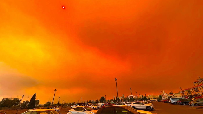 An Orange sky caused by flames from a forest fire approaching a car park filled with cars