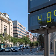 A digital street thermometer in Seville displays 48°C during an extreme heatwave as pedestrians cross the road under clear blue skies.