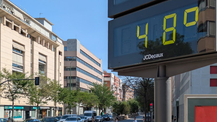 A digital street thermometer in Seville displays 48°C during an extreme heatwave as pedestrians cross the road under clear blue skies.
