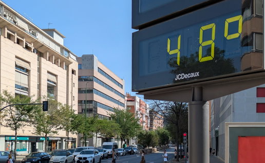 A digital street thermometer in Seville displays 48°C during an extreme heatwave as pedestrians cross the road under clear blue skies.