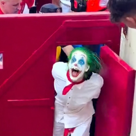 Person dressed as the Joker with green hair and white face paint being escorted from the Pamplona bullring during San Fermín.