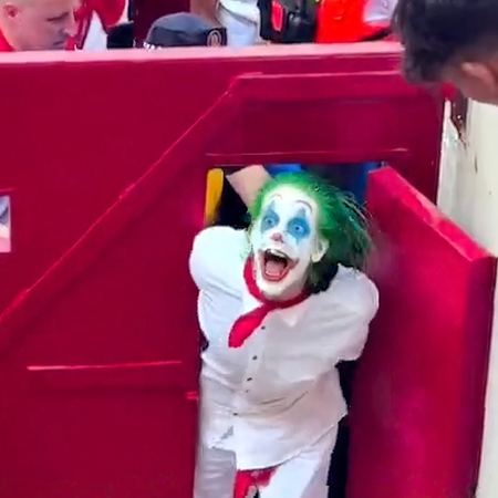 Person dressed as the Joker with green hair and white face paint being escorted from the Pamplona bullring during San Fermín.
