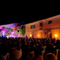 A seated crowd watching an outdoor night-time concert between traditional buildings in Oria.