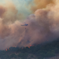 A wildfire spreading through a rural area with a helicopter dropping water on flames