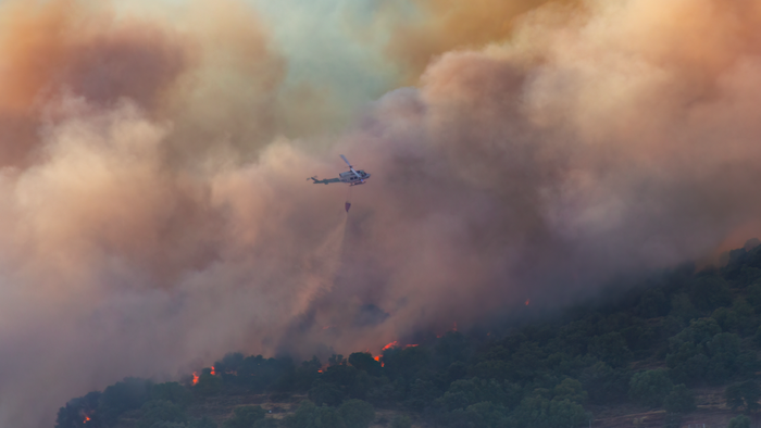 A wildfire spreading through a rural area with a helicopter dropping water on flames