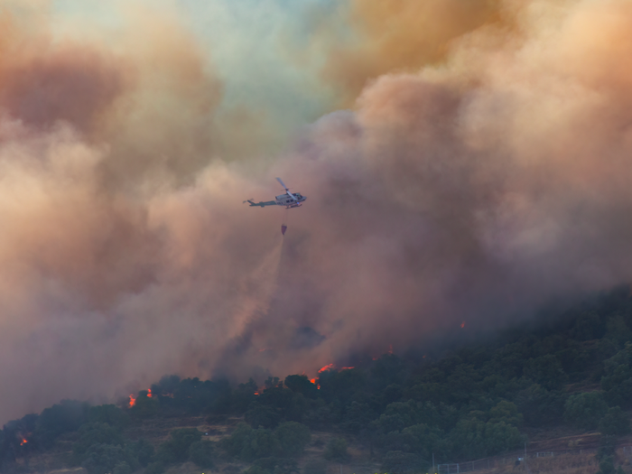 A wildfire spreading through a rural area with a helicopter dropping water on flames