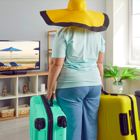 Woman at home with suitcases and sunhat looking at a beach on a tv screen