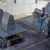 Emergency responders and firefighters gathered around a large metal underground rubbish chute on a paved outdoor site, with one civilian man in a white t-shirt observing.