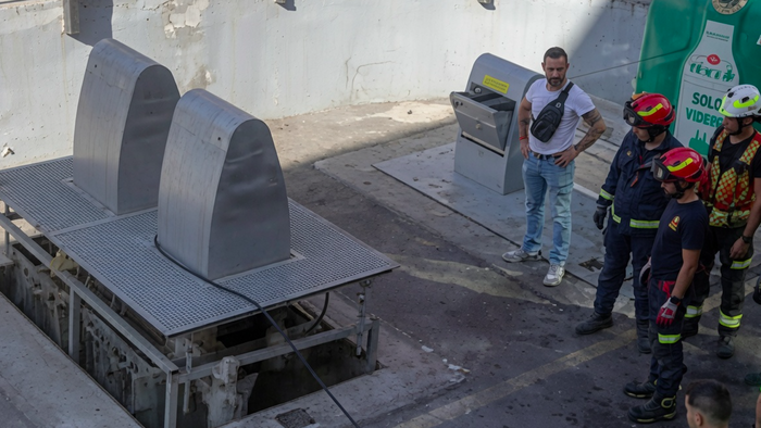 Emergency responders and firefighters gathered around a large metal underground rubbish chute on a paved outdoor site, with one civilian man in a white t-shirt observing.